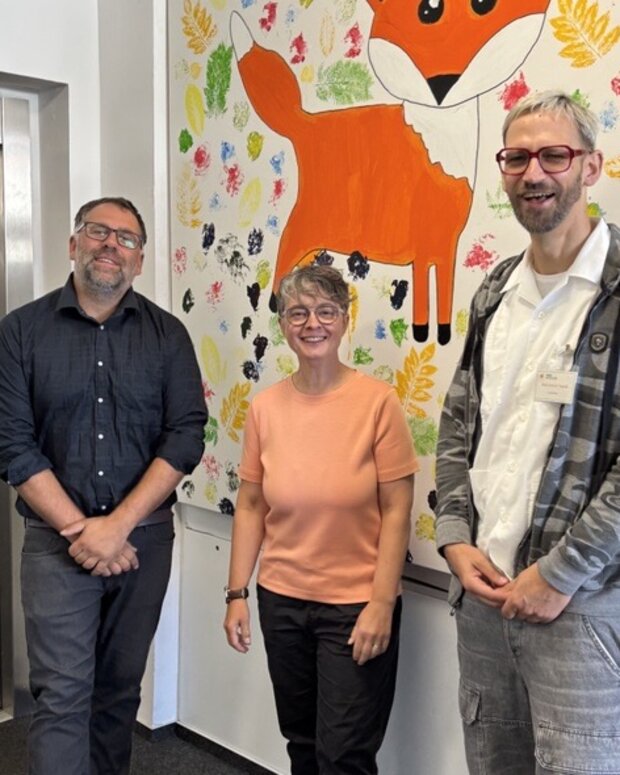 Drei Personen stehen in einem Flur der WBS SCHULEN Berlin neben einem Aufzug vor einem großen, farbenfrohen Wandbild mit einem orangefarbenen Fuchs. Der Hintergrund des Bildes ist mit bunten Blatt- und Farbabdrucken gestaltet.