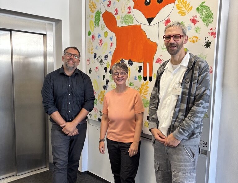 Drei Personen stehen in einem Flur der WBS SCHULEN Berlin neben einem Aufzug vor einem großen, farbenfrohen Wandbild mit einem orangefarbenen Fuchs. Der Hintergrund des Bildes ist mit bunten Blatt- und Farbabdrucken gestaltet.
