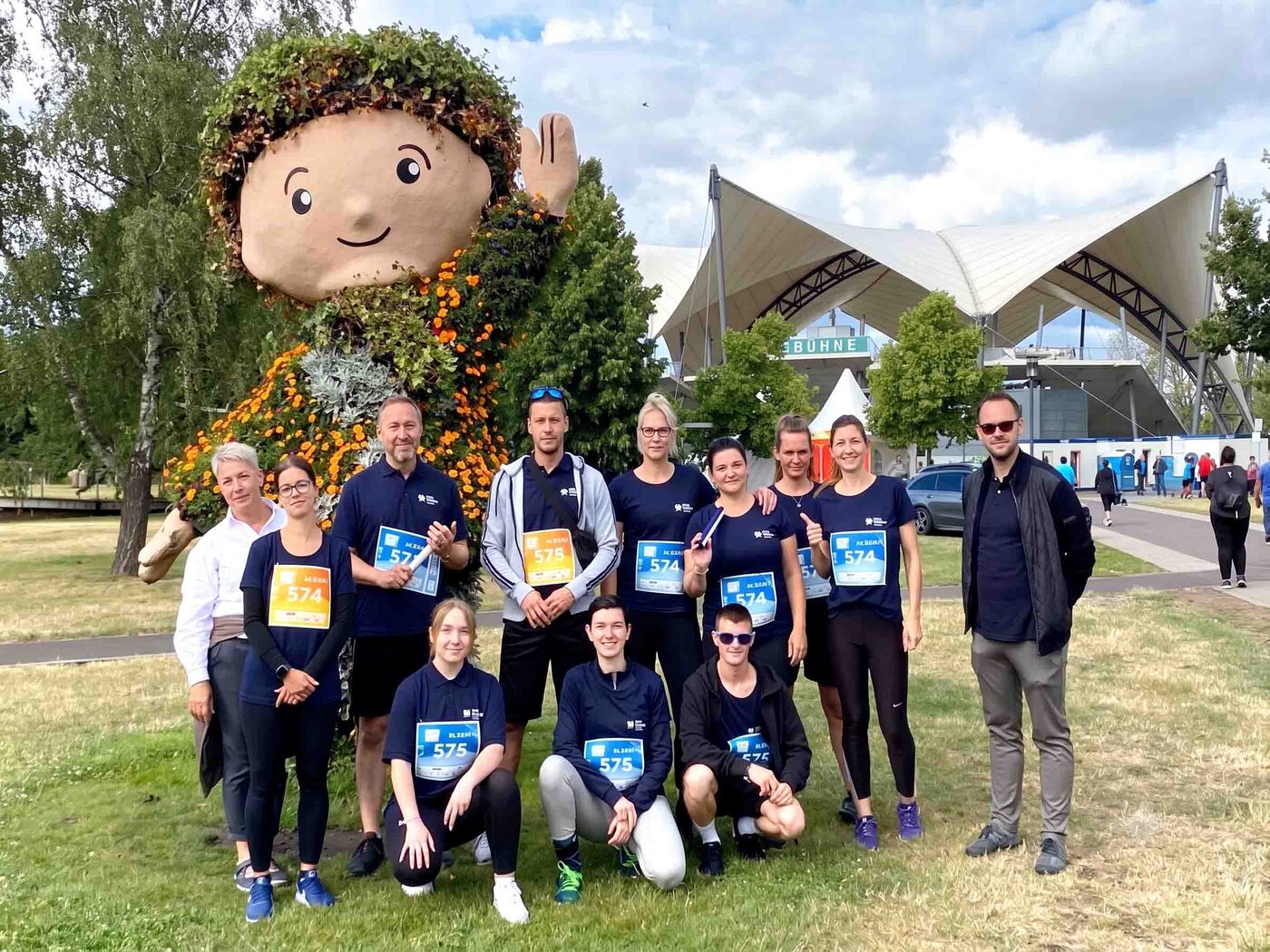Team des Standorts WBS SCHULEN Magdeburg beim Firmenlauf. Die Teilnehmenden posieren mit Startnummern vor einer großen Blumenfigur und einem Stadiongelände im Hintergrund.