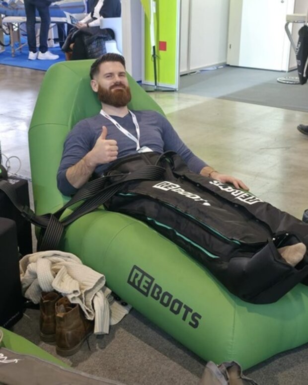 Eine Lehrkraft für Physiotherapie-Ausbildungen testet an einem Messestand ein REBOOTS-Kompressionsmassage-System auf einer grünen Liege.