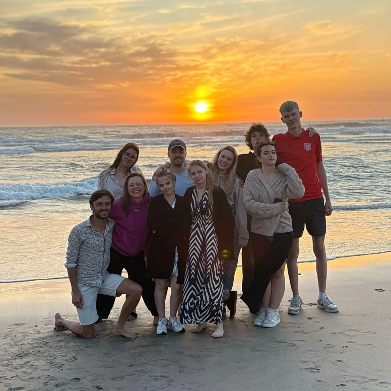 Mehrere Teilnehmende eines Auslandspraktikums stehen für ein Gruppenfoto am Strand von Barcelona. Im Hintergrund sind das Meer und ein farbenreicher Sonnenuntergang zu sehen, der den Himmel in warmes Abendlicht taucht.