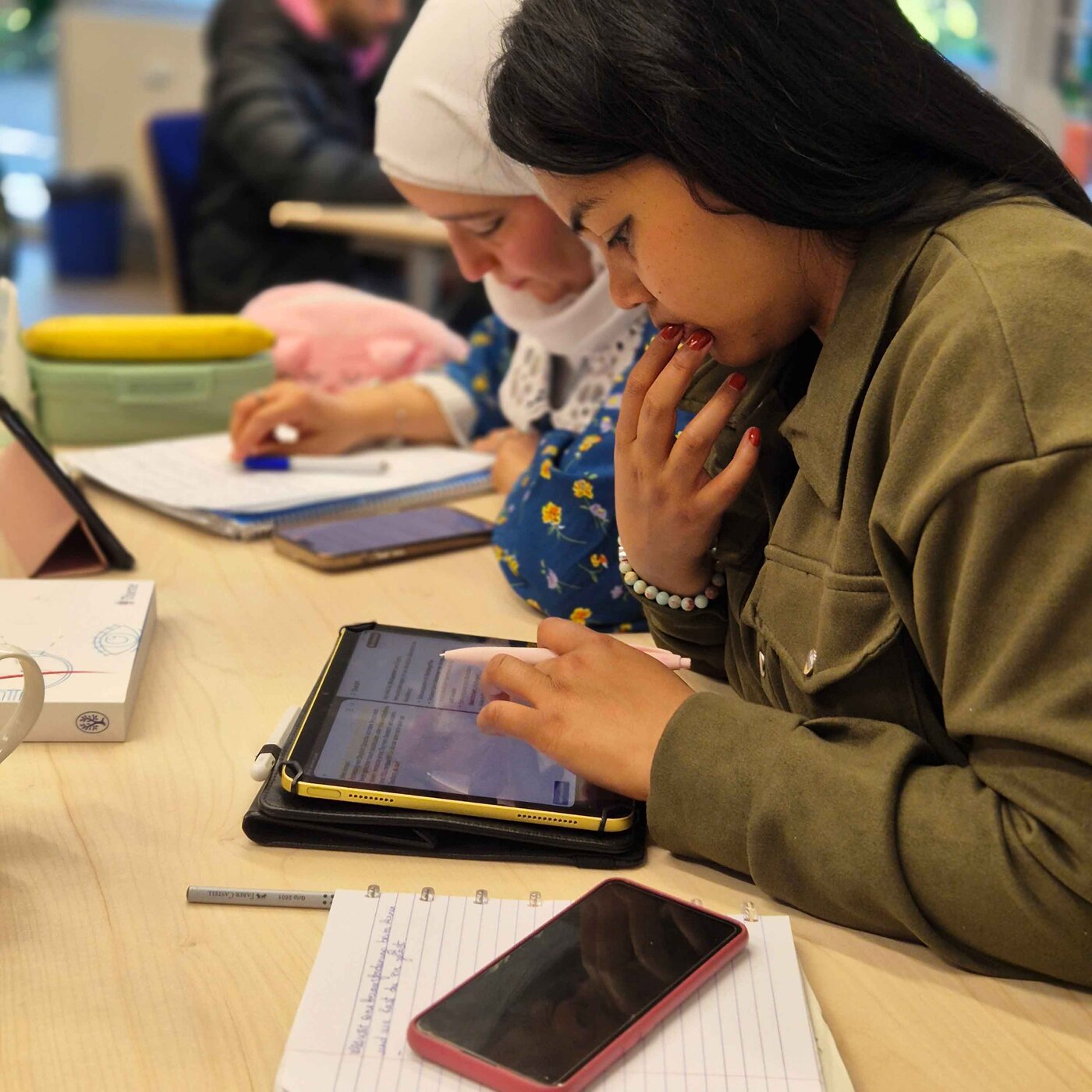 Zwei Lernende sitzen an einem Tisch im Rahmen des Unterrichtskonmzepts SOL (selbstorganisiertes Lernen). Eine Person liest auf einem Tablet mit Eingabestift, die andere macht handschriftliche Notizen. Vor ihnen liegen Lernmaterialien und ein Smartphone.