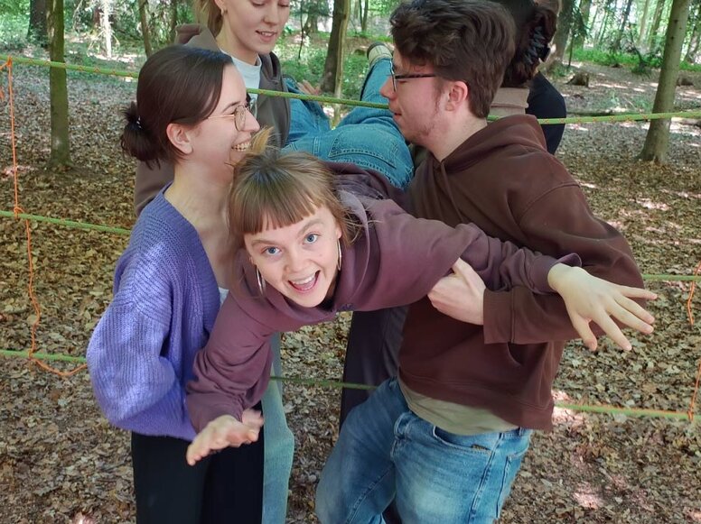 Mehrere Schüler:innen stehen auf einer Waldlichtung innerhalb eines aus Seilen gebildeten Gitters und unterstützen sich gegenseitig dabei, eine Person durch eine Öffnung im Seilnetz zu bewegen. Die Szene zeigt eine kooperative Teamübung im Freien.