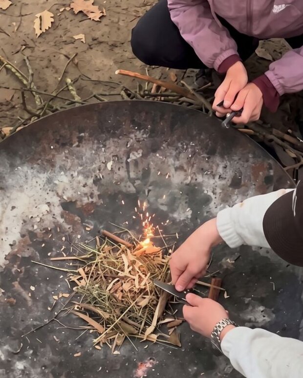 Drei Personen entzünden gemeinsam ein kleines Feuer in einer großen Metallschale im Freien. Um die Feuerstelle liegen Äste und Laub, im Hintergrund stehen Biertischgarnituren.