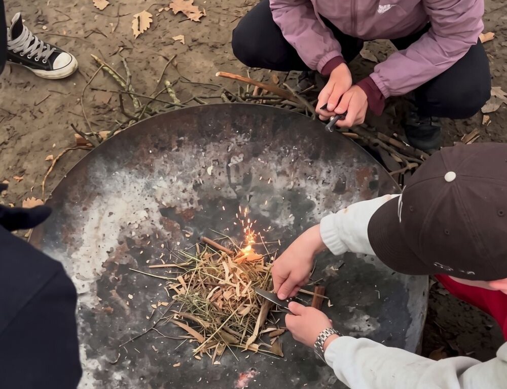Drei Personen entzünden gemeinsam ein kleines Feuer in einer großen Metallschale im Freien. Um die Feuerstelle liegen Äste und Laub, im Hintergrund stehen Biertischgarnituren.