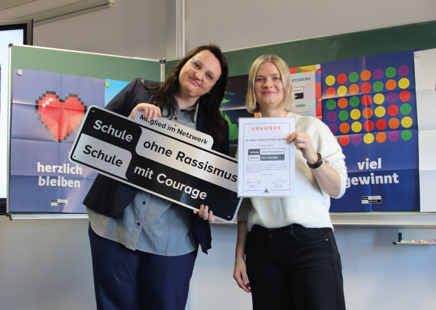 Zwei Frauen halten das Schild „Schule ohne Rassismus – Schule mit Courage“ hoch. Sie stehen vor einer Tafel mit Plakaten zum Thema Vielfalt und lächeln.