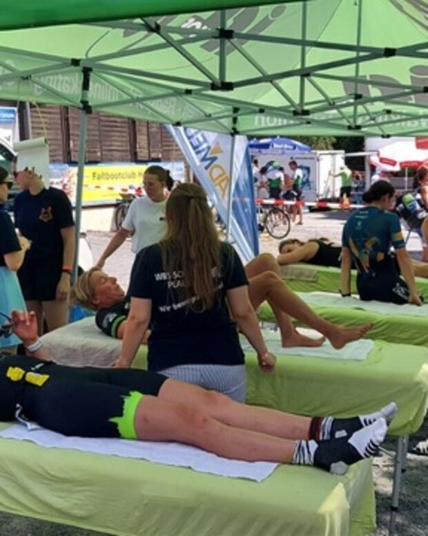 Mehrere Behandlungsliegen unter einem grünen Pavillonzelt bei einer Sportveranstaltung im Freien. Auf den Liegen werden Teilnehmende von mehreren Physiotherapeut:innenbetreut.