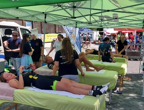 Mehrere Behandlungsliegen unter einem grünen Pavillonzelt bei einer Sportveranstaltung im Freien. Auf den Liegen werden Teilnehmende von mehreren Physiotherapeut:innenbetreut.
