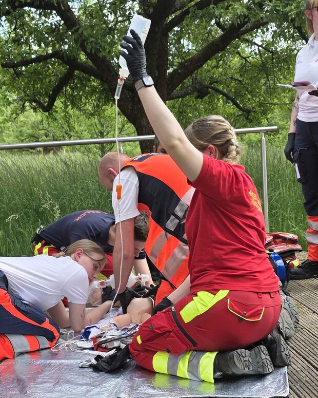 Mehrere Rettungsdienst-Auszubildende üben auf einer Holzterrasse im Freien einen Notfalleinsatz an einer Übungspuppe. Eine Person hält einen Infusionsbeutel, während andere die simulierte Versorgung durchführen; im Hintergrund stehen Bäume und hohes Gras.