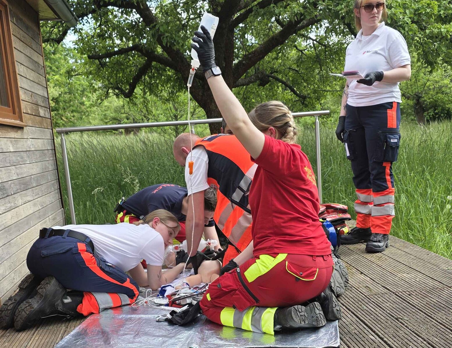 Mehrere Rettungsdienst-Auszubildende üben auf einer Holzterrasse im Freien einen Notfalleinsatz an einer Übungspuppe. Eine Person hält einen Infusionsbeutel, während andere die simulierte Versorgung durchführen; im Hintergrund stehen Bäume und hohes Gras.