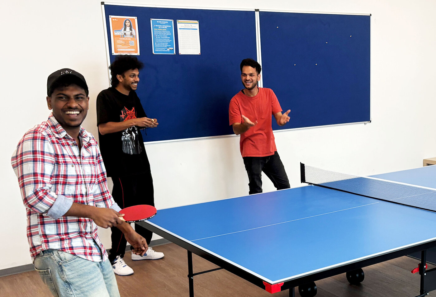 Pause bei den WBS SCHULEN in Troisdorf: Drei junge Männer stehen lachend an einer Tischtennisplatte in einem hellen Pausenraum und spielen.