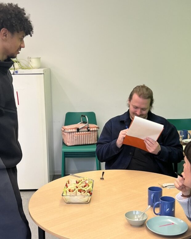 Schüler und Juroren in einer Gemeinschaftsküche mit selbst zubereiteten Snacks auf einem runden Tisch.