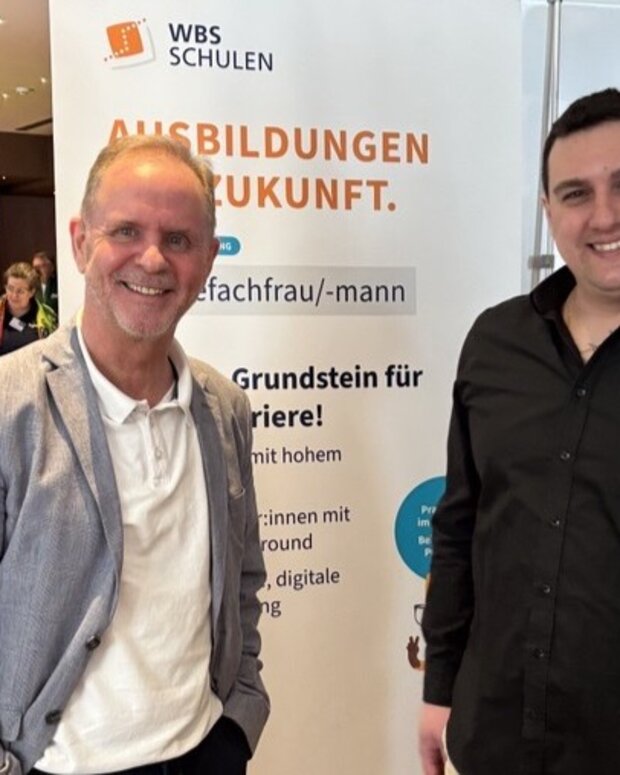 Zwei Personen stehen an einem Messestand der WBS SCHULEN in einer Veranstaltungshalle. Im Hintergrund ist ein Banner mit dem Schriftzug „AUSBILDUNGEN MIT ZUKUNFT“ sowie Informationen zur Ausbildung als Pflegefachfrau/-mann zu sehen.