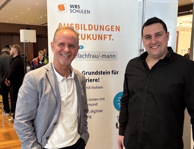 Zwei Personen stehen an einem Messestand der WBS SCHULEN in einer Veranstaltungshalle. Im Hintergrund ist ein Banner mit dem Schriftzug „AUSBILDUNGEN MIT ZUKUNFT“ sowie Informationen zur Ausbildung als Pflegefachfrau/-mann zu sehen.