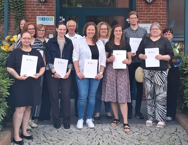 Gruppenfoto der Absolventinnen und Absolventen einer Weiterbildung zur Praxisanleitung mit ihren Abschlussurkunden vor dem Schulgebäude.