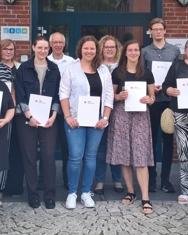 Gruppenfoto der Absolventinnen und Absolventen einer Weiterbildung zur Praxisanleitung mit ihren Abschlussurkunden vor dem Schulgebäude.
