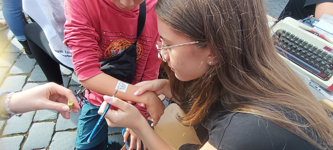 Mehrere Schülerinnen und Schüler gestalten an einem Stand im Freien temporäre Tattoos auf einem Unterarm. Im Vordergrund wird mit einem Pinsel ein Motiv aufgetragen, im Hintergrund ist eine Schreibmaschine auf dem Tisch zu sehen.