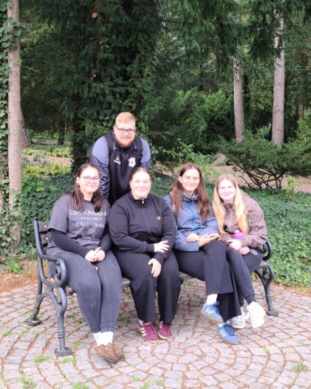 Gruppenfoto von fünf Schüler:innen auf und an einer Parkbank auf dem Westfriedhof Magdeburg vor dichtem Baumbestand in einer Parkanlage.