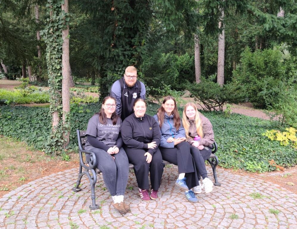 Gruppenfoto von fünf Schüler:innen auf und an einer Parkbank auf dem Westfriedhof Magdeburg vor dichtem Baumbestand in einer Parkanlage.