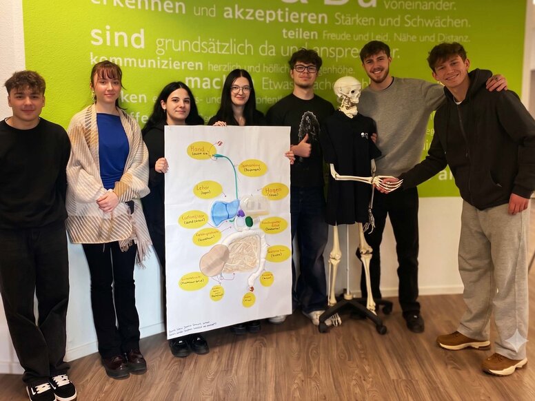 Fünf Auszubildende der Pflege präsentieren stolz ein selbstgemaltes, detailliertes Plakat des menschlichen Verdauungssystems. Rechts im Bild schüttelt ein Schüler lachend die Hand eines Skelett-Modells.