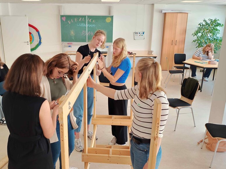 Mehrere Schüler:innen arbeiten gemeinsam in einem Gruppenraum an einem Holzgestell. Während einige Bauteile montieren und ausrichten, sitzen weitere Teilnehmende im Raum an Tischen oder beobachten die Aufgabe.