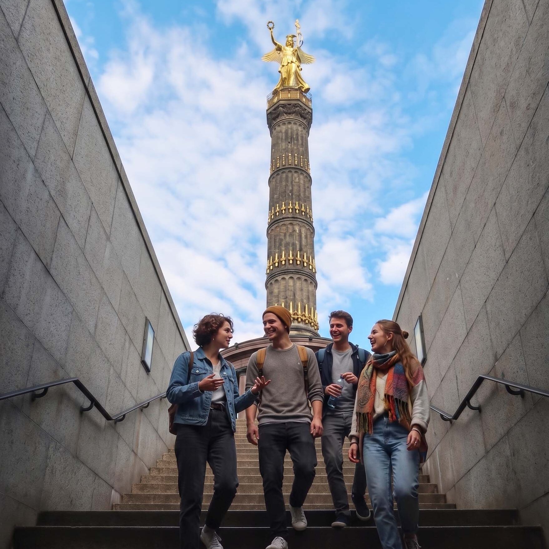 Vier junge Erwachsene gehen fröhlich eine Steintreppe hinauf, mit der Berliner Siegessäule und der Goldelse-Statue im Hintergrund vor blauem Himmel.