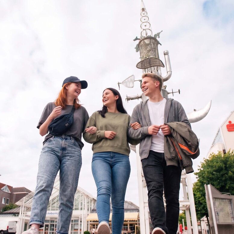 Drei lachende junge Menschen gehen Arm in Arm über den Marktplatz in Aurich. Im Hintergrund ragt der Sous Turm in den Himmel.