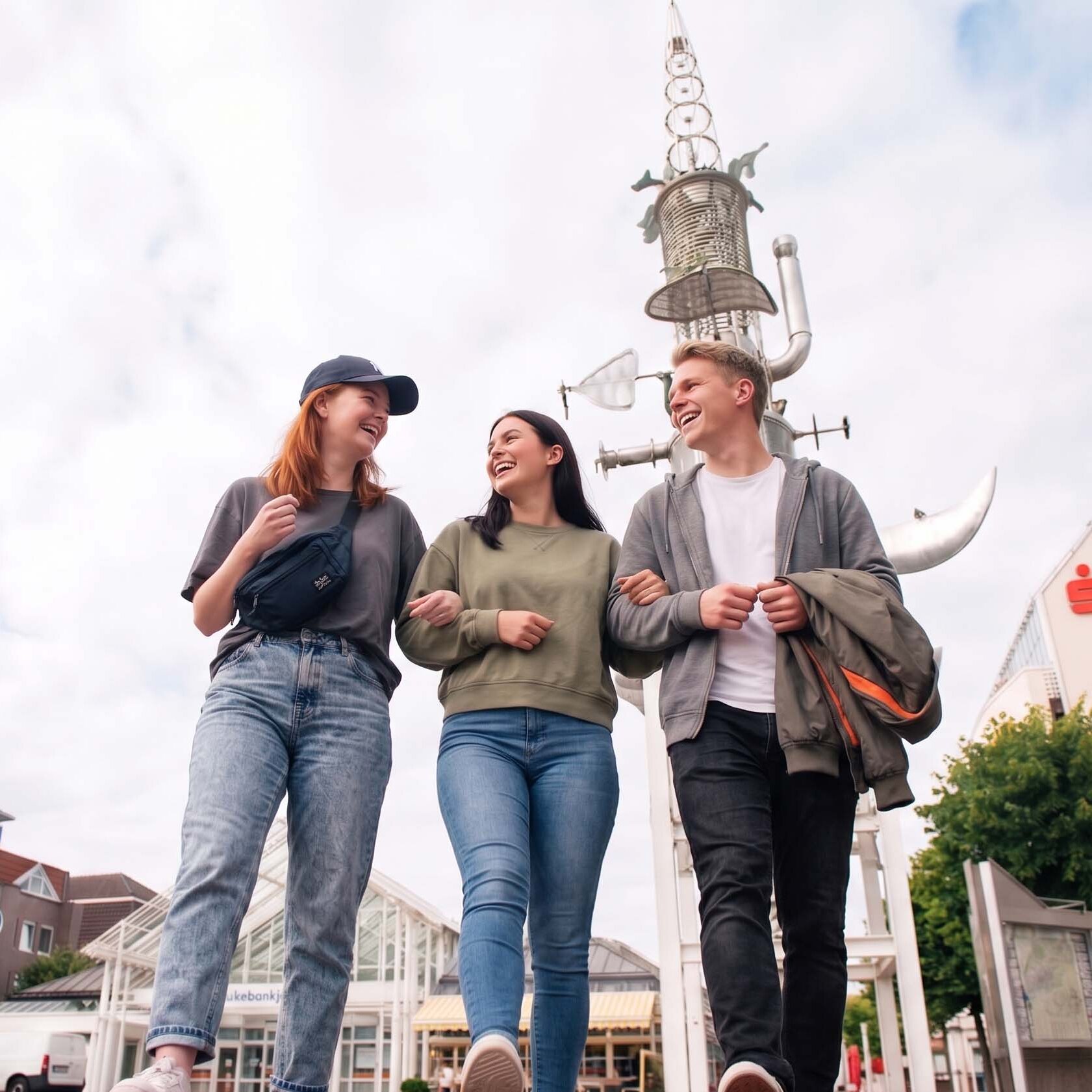 Drei lachende junge Menschen gehen Arm in Arm über den Marktplatz in Aurich. Im Hintergrund ragt der Sous Turm in den Himmel.