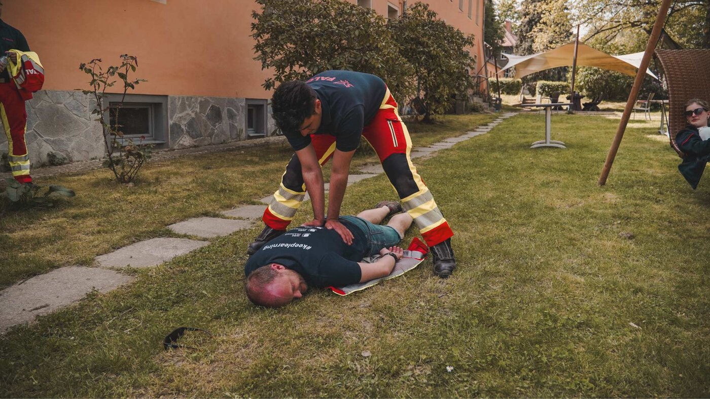 Eine Person in Rettungsdienstkleidung übt auf einer Wiese die Versorgung einer am Boden liegenden Person im Rahmen eines Praxistrainings.