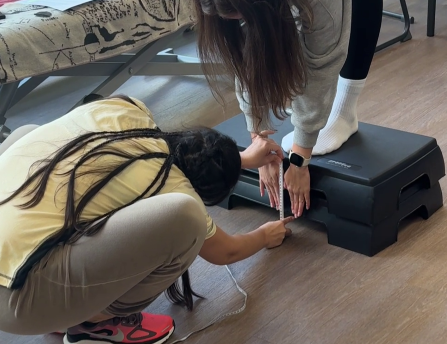 Zwei Personen messen in einem Therapieraum die Beweglichkeit eines ausgestreckten Fußes auf einer erhöhten Plattform mit einem Maßband. Im Hintergrund ist eine Behandlungsliege zu sehen.