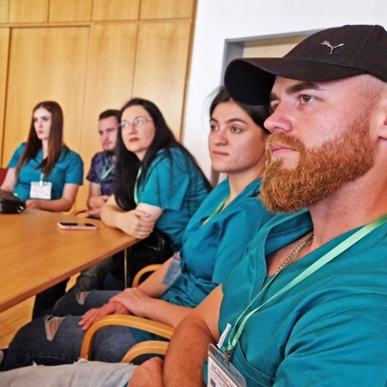 Mehrere Lernende in grünen Kasacks sitzen an einem Holztisch in einem Seminarraum und blicken konzentriert aus dem Bildrand heraus.