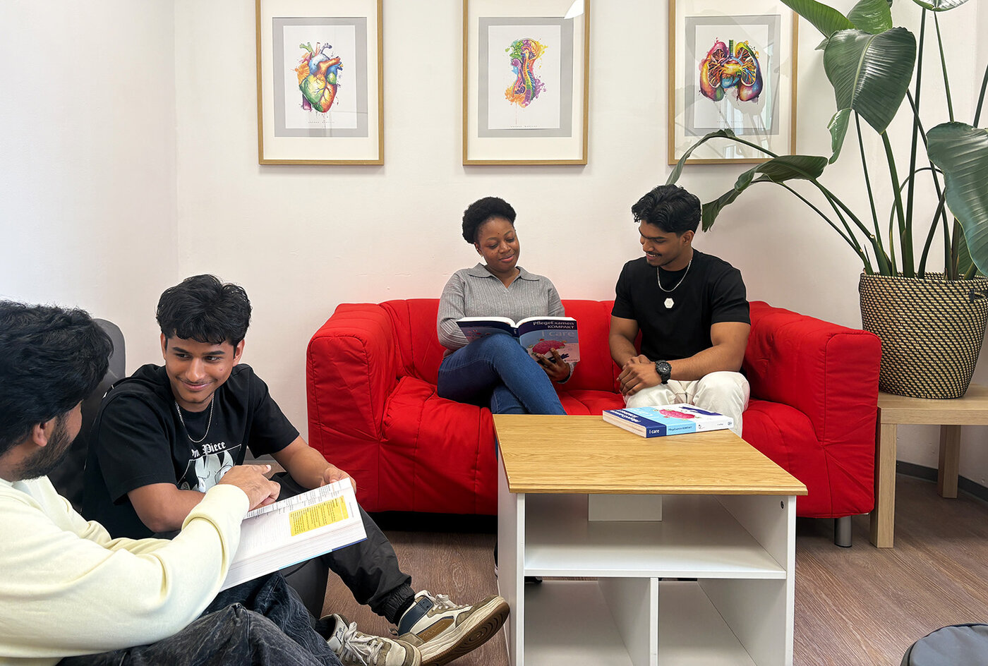 Vier Lernende sitzen in einem Aufenthaltsraum mit rotem Sofa und lernen gemeinsam aus Fachbüchern. An der Wand hängen bunte anatomische Bilder.
