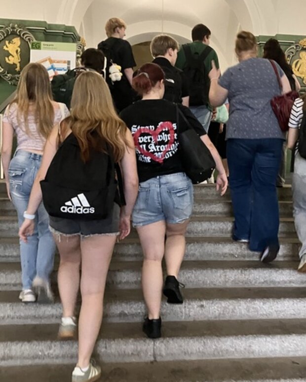 Eine Gruppe von Lernenden und begleitenden Personen geht eine breite Steintreppe in einem historischen Gebäude hinauf. Der Innenraum ist durch grüne Wände, Rundbögen und dekorative Wappen an den Wänden geprägt.