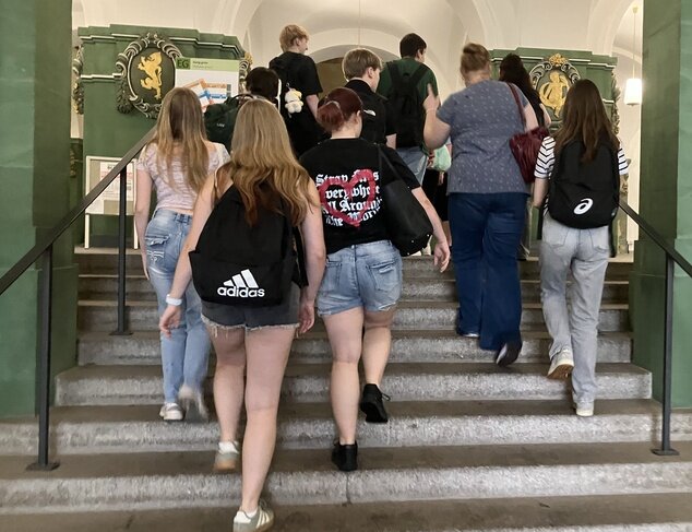 Eine Gruppe von Lernenden und begleitenden Personen geht eine breite Steintreppe in einem historischen Gebäude hinauf. Der Innenraum ist durch grüne Wände, Rundbögen und dekorative Wappen an den Wänden geprägt.