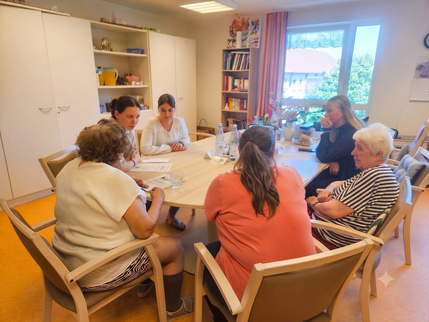 Auszubildende und Senioren sitzen gemeinsam an einem runden Tisch in einem Aufenthaltsraum eines Seniorenwohnheims. Sie tauschen sich anhand von Unterlagen aus. Im Raum stehen Schränke, Regale und Getränke auf dem Tisch, Tageslicht fällt durch ein großes Fenster.