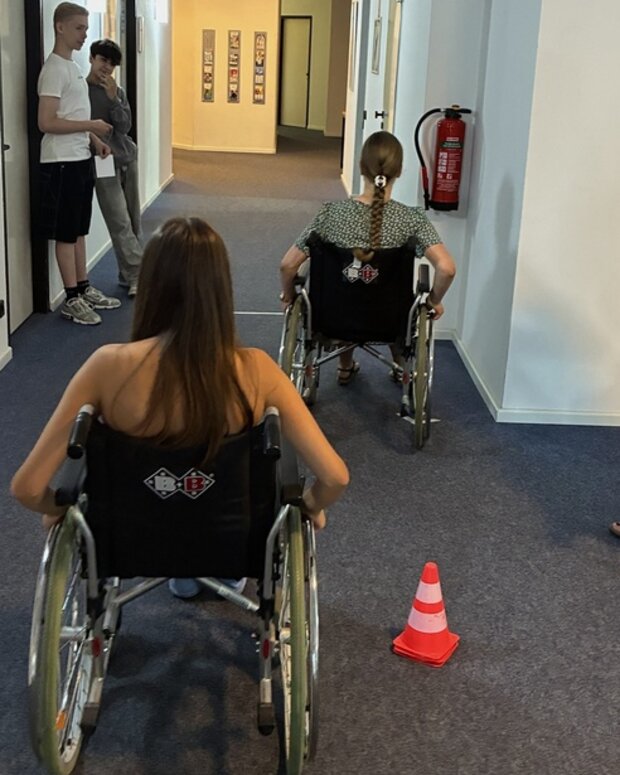 Rollstuhlparcours in einem Flur: Zwei Schülerinnen üben das Manövrieren von Rollstühlen, beobachtet von weiteren Teilnehmenden und der Übungsleiterin.