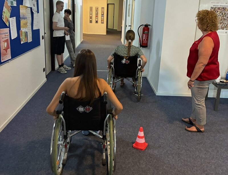 Rollstuhlparcours in einem Flur: Zwei Schülerinnen üben das Manövrieren von Rollstühlen, beobachtet von weiteren Teilnehmenden und der Übungsleiterin.