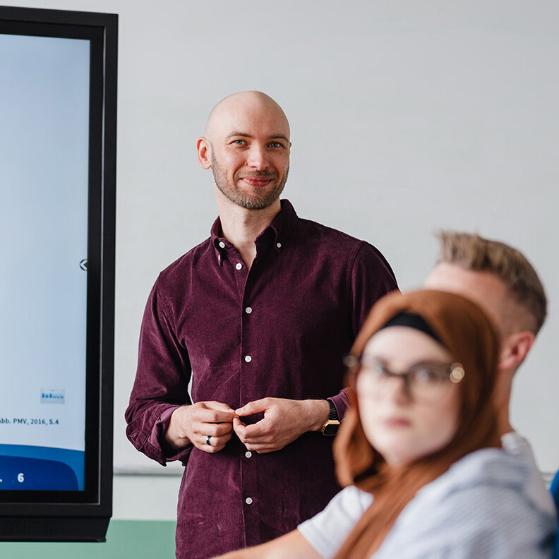 Ein Lernbegleiter steht neben dem digitalen Whiteboard und hört der Antwort einer Schülerin zu