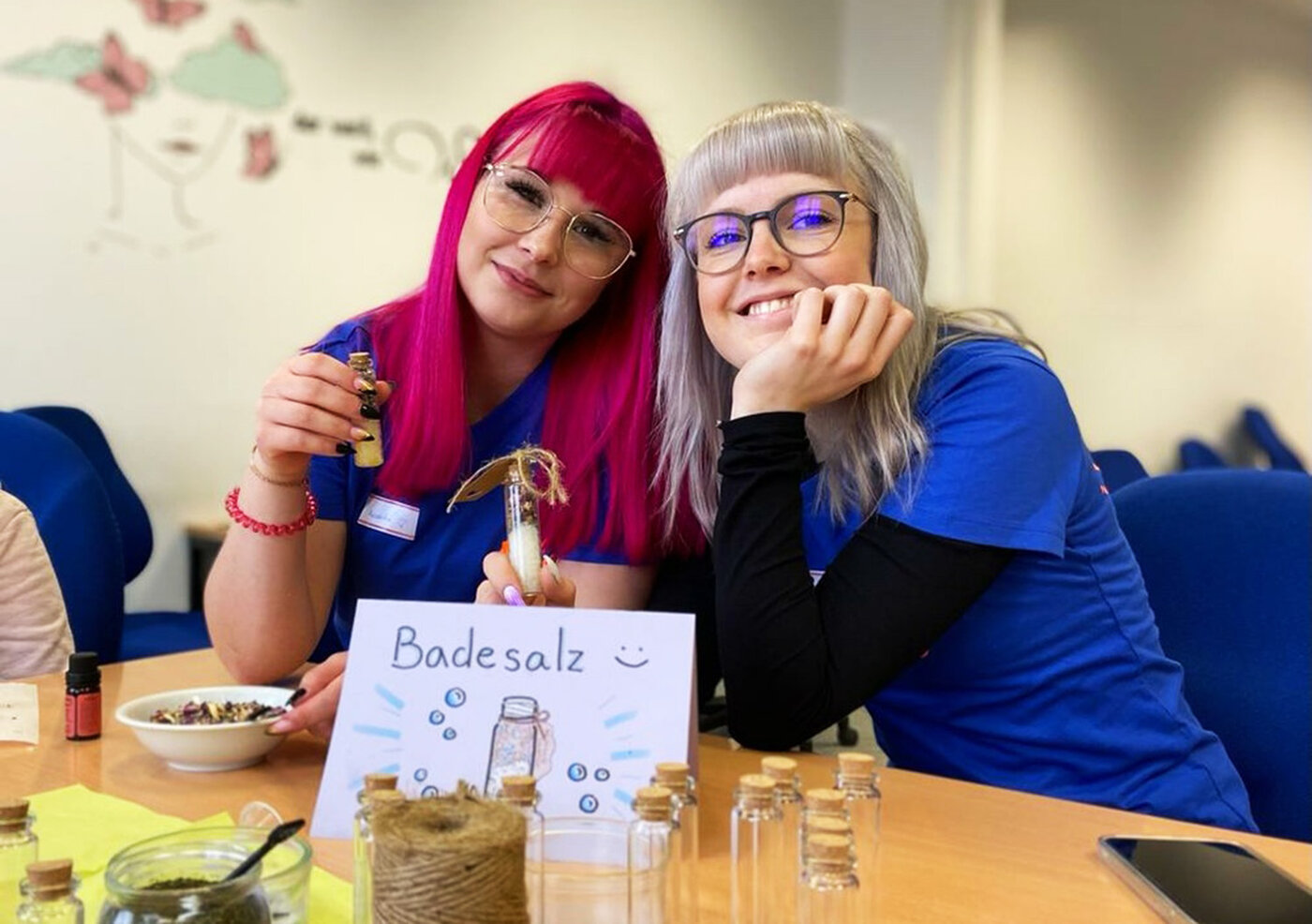 Zwei lächelnde Auszubildende zur Pflegefachkraft sitzen an einem Tisch. Eine von ihnen hält während eines Workshops ein mit Badesalz gefülltes Glasfläschchen in der Hand. 