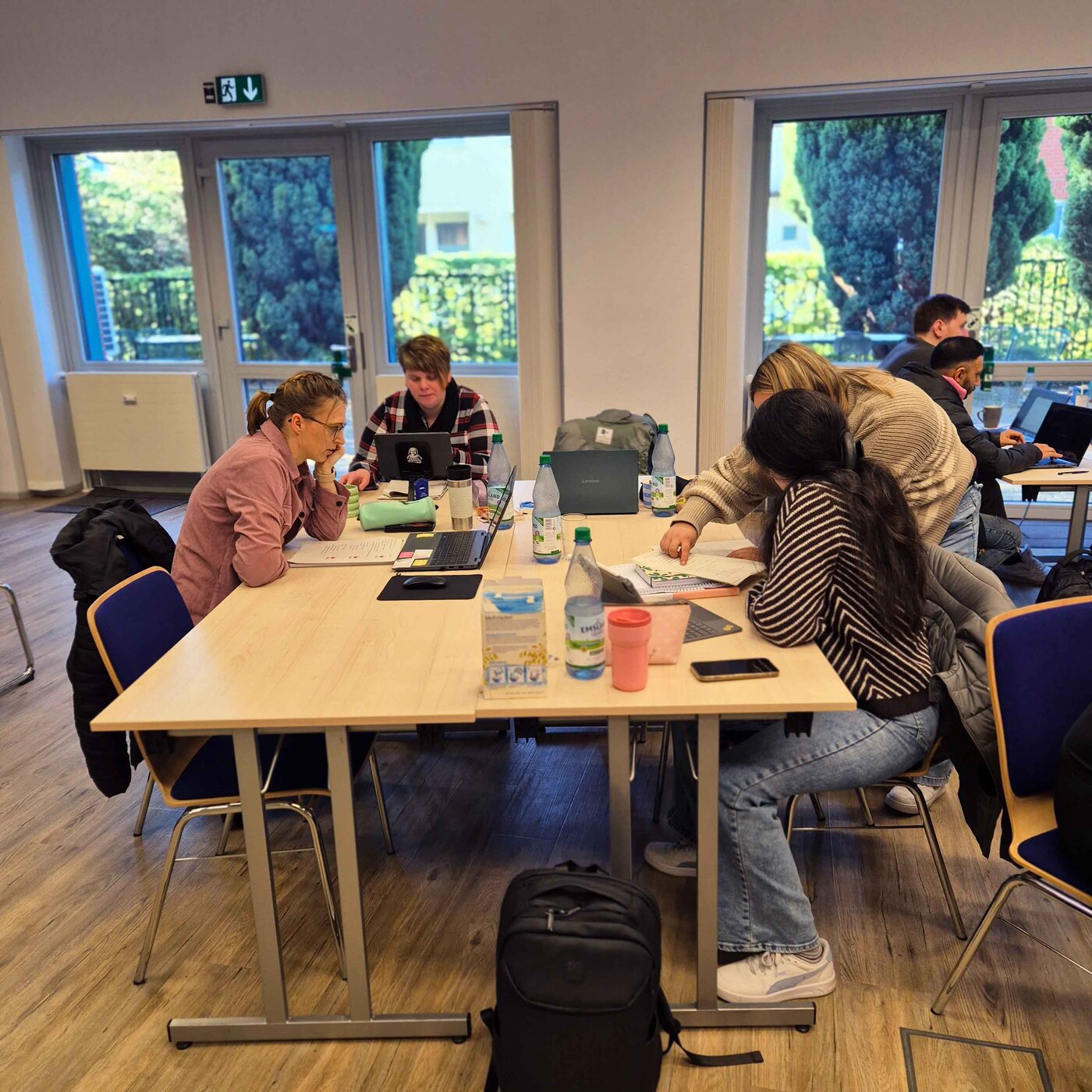 Mehrere Personen sitzen in einem hellen Unterrichtsraum an Tischen und arbeiten in Gruppen im Rahmen des Unterrichtskonzepts SOL (selbstorganisiertes Lernen). Sie lernen gemeinsam mithilfe von Laptops und gedruckten Unterlagen.