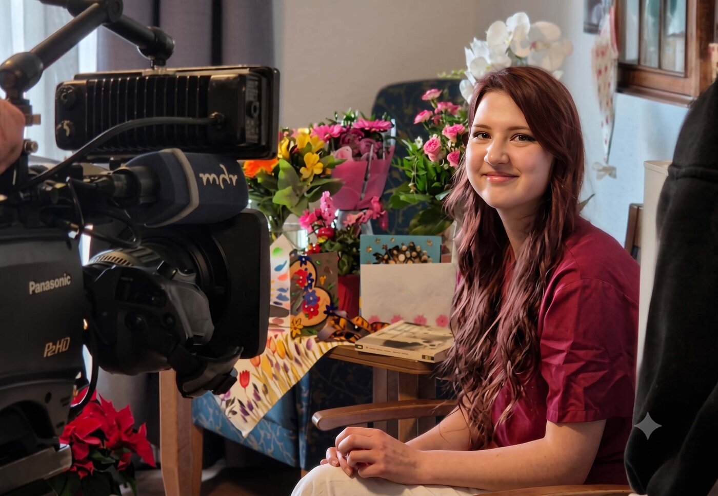 Eine junge, lächelnde Auszubildende mit langen rotbraunen Haaren sitzt auf einem Stuhl und wird von einer professionellen MDR-Fernsehkamera gefilmt. Neben ihr steht ein Tisch, der reichlich mit bunten Blumensträußen, Grußkarten und Geschenken geschmückt ist.