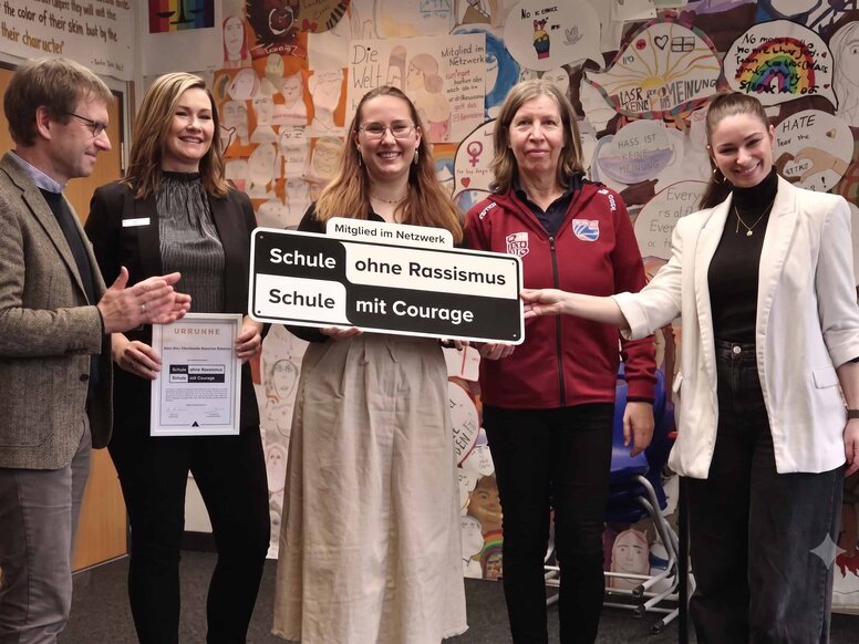 Fünf Personen präsentieren lächelnd ein Schild mit der Aufschrift „Schule ohne Rassismus – Schule mit Courage“ und eine Urkunde. Die Wand im Hintergrund ist mit vielen bunten Anti-Rassismus-Plakaten beklebt.