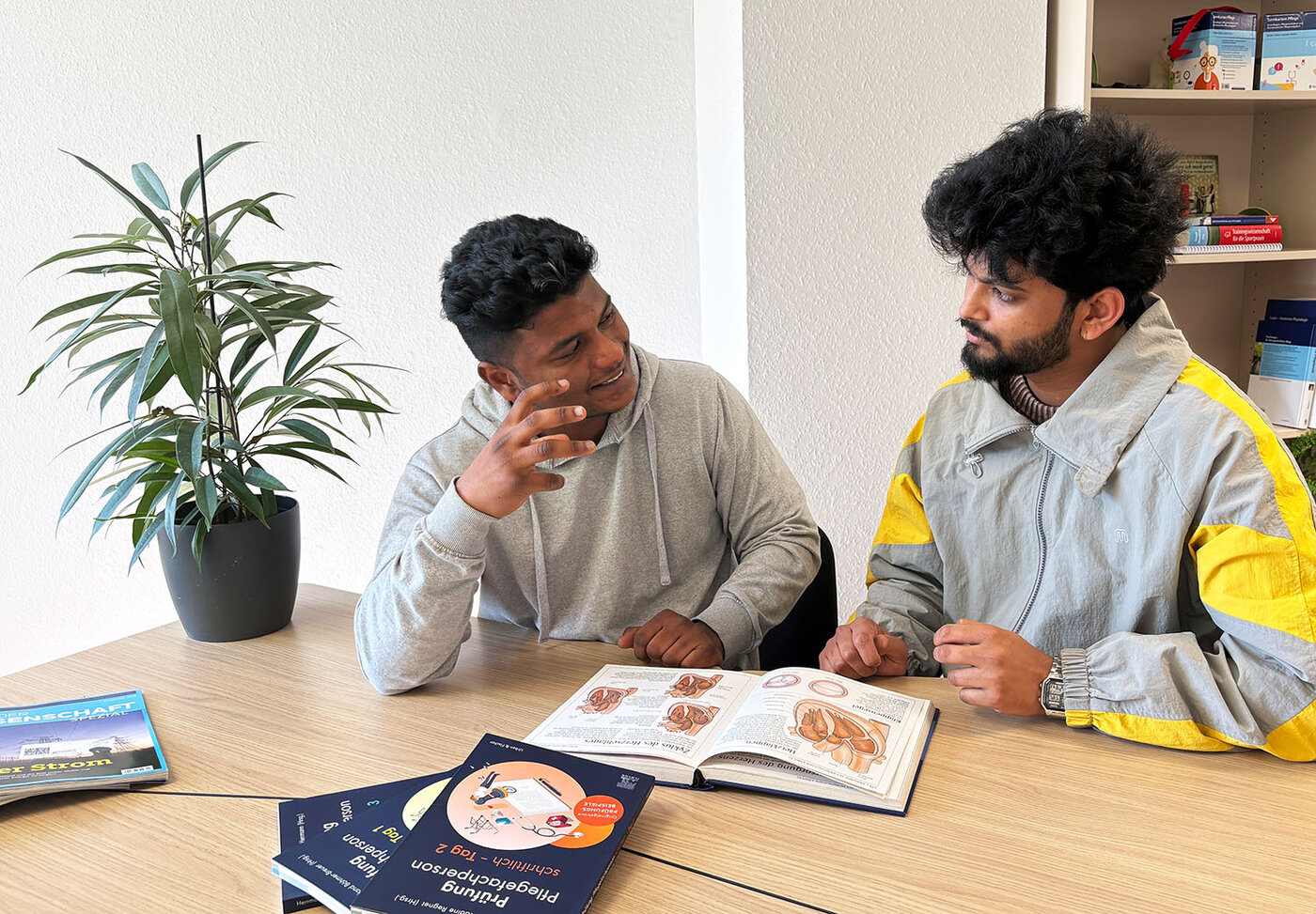 Zwei junge Männer sitzen an einem Tisch und lernen gemeinsam. Vor ihnen liegen aufgeschlagene Fachbücher mit anatomischen Abbildungen.