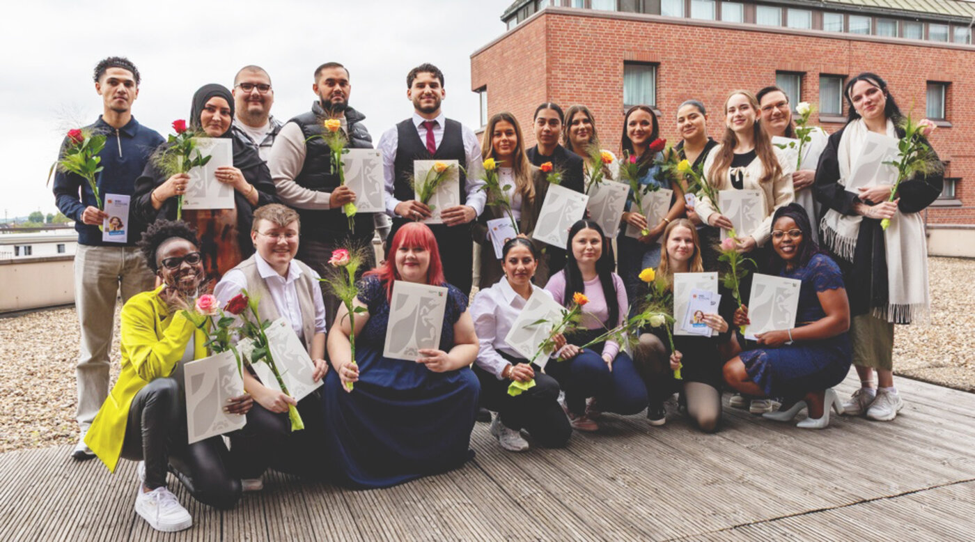 Eine große, feierliche Gruppe von Absolventinnen und Absolventen posiert auf der Dachterrasse des Schulgebäudes der WBS SCHULEN Hamm. Alle halten stolz ihre Abschlusszeugnisse und bunte Rosen in den Händen und lächeln in die Kamera.
