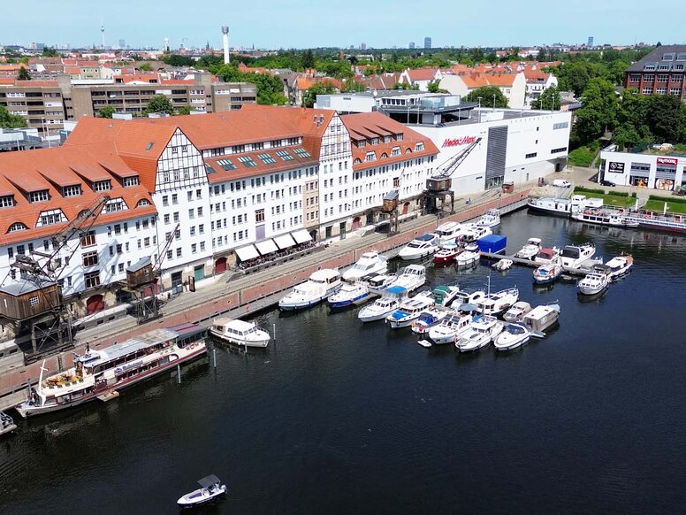 Luftaufnahme des Lorenzweg 5 in Berlin.Im Vordergrund liegt Wasser, auf dem mehrere Boote und ein Ausflugsschiff zu sehen sind. Am Ufer steht ein langes, mehrstöckiges Backsteingebäude mit roten Ziegeldächern, weißen Fassaden und großen Kränen. Im Hintergrund ist eine weitläufige Stadtlandschaft erkennbar.