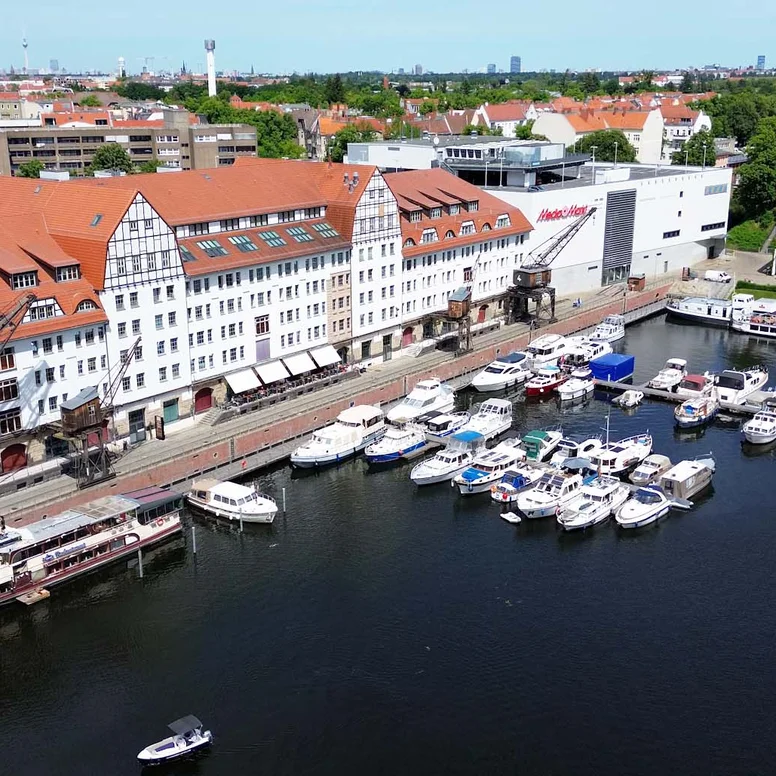 Luftaufnahme des Lorenzweg 5 in Berlin.Im Vordergrund liegt Wasser, auf dem mehrere Boote und ein Ausflugsschiff zu sehen sind. Am Ufer steht ein langes, mehrstöckiges Backsteingebäude mit roten Ziegeldächern, weißen Fassaden und großen Kränen. Im Hintergrund ist eine weitläufige Stadtlandschaft erkennbar.