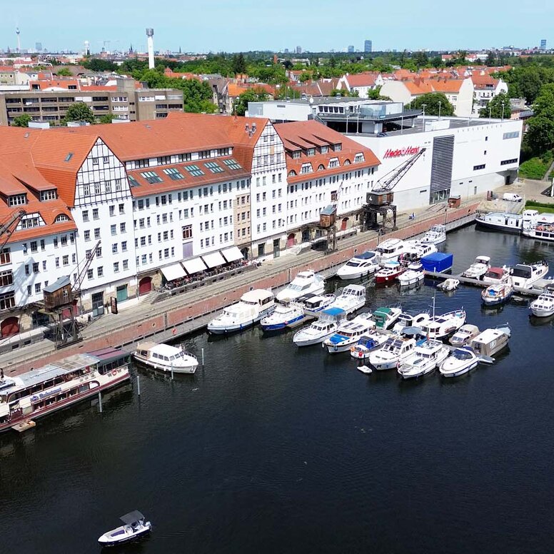 Luftaufnahme des Lorenzweg 5 in Berlin.Im Vordergrund liegt Wasser, auf dem mehrere Boote und ein Ausflugsschiff zu sehen sind. Am Ufer steht ein langes, mehrstöckiges Backsteingebäude mit roten Ziegeldächern, weißen Fassaden und großen Kränen. Im Hintergrund ist eine weitläufige Stadtlandschaft erkennbar.