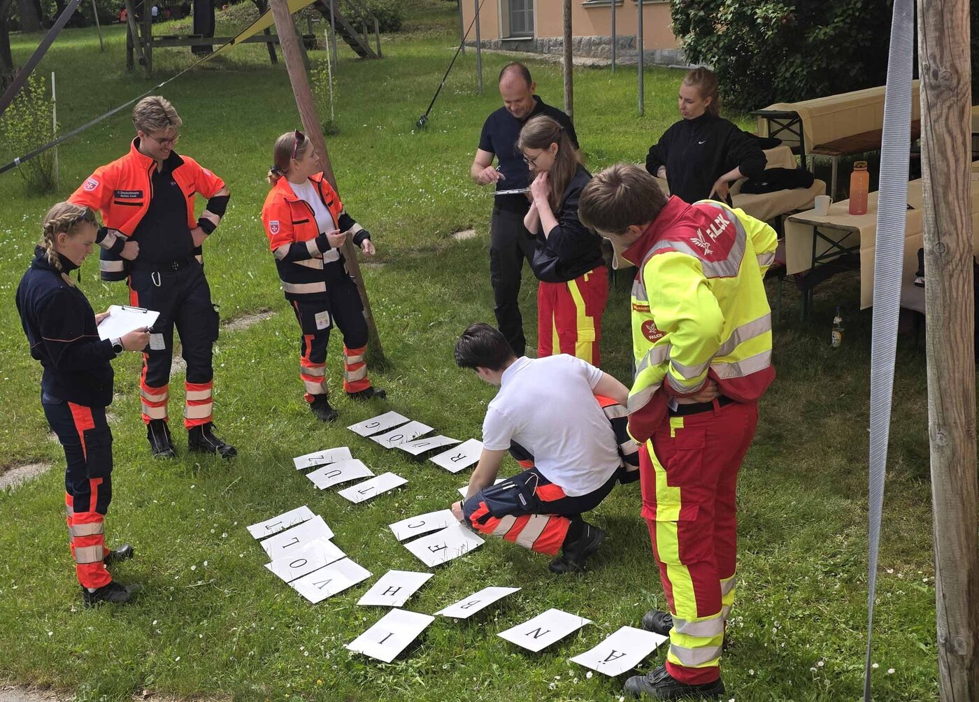 Mehrere Personen in Rettungsdienstkleidung stehen bei einer Ausbildungsübung im Freien um auf dem Rasen ausgelegte Buchstabenkarten und lösen gemeinsam eine Aufgabe.