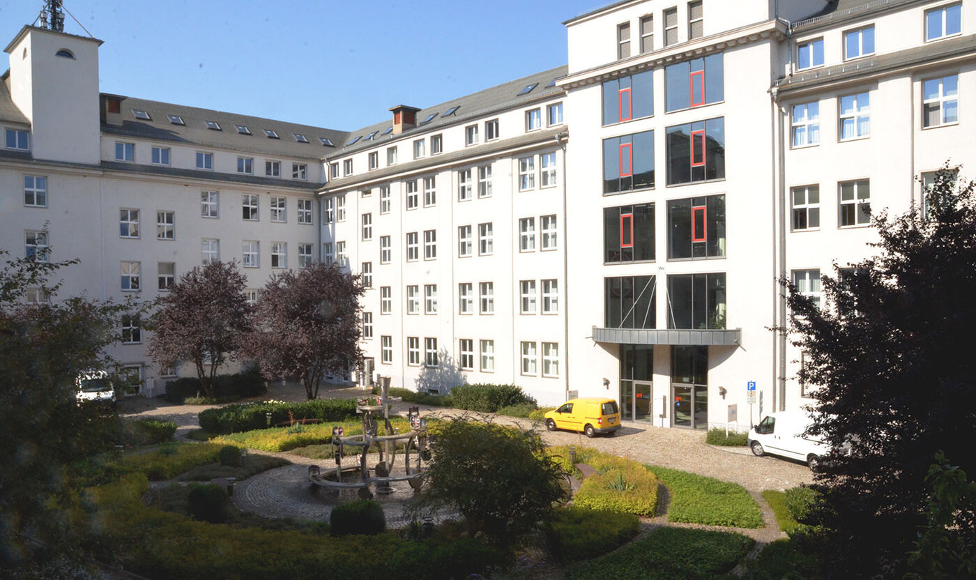 Die Außenansicht des Schulgebäudes in der Kauffahrtei 25 in 09120 Chemnitz. Das Gebäude ist U-förmig gebaut und umschließt einen gepflasterten Innenhof. Im Hof befinden sich Pflanzen, Bäume und ein Springbrunnen. Ein gelbes und ein weißes Auto parken vor dem verglasten Eingangsbereich.