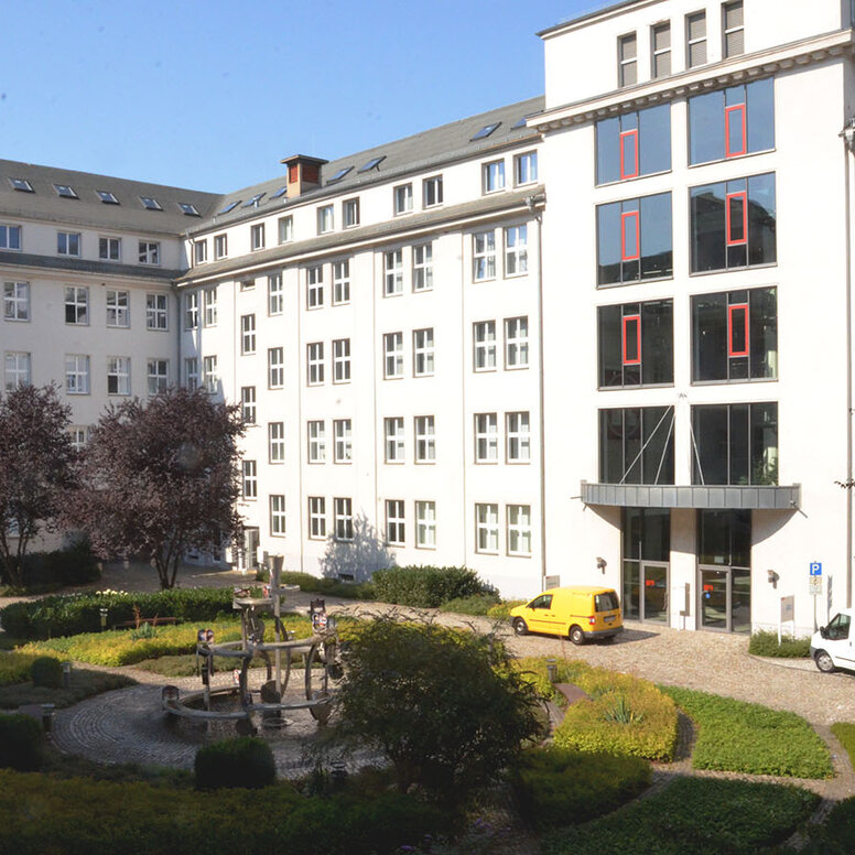 Die Außenansicht des Schulgebäudes in der Kauffahrtei 25 in 09120 Chemnitz. Das Gebäude ist U-förmig gebaut und umschließt einen gepflasterten Innenhof. Im Hof befinden sich Pflanzen, Bäume und ein Springbrunnen. Ein gelbes und ein weißes Auto parken vor dem verglasten Eingangsbereich.